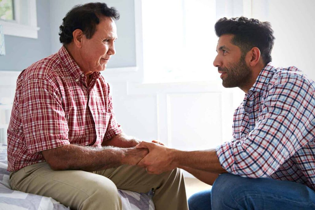 Senior man seated with adult man kneeling beside him holding his hand